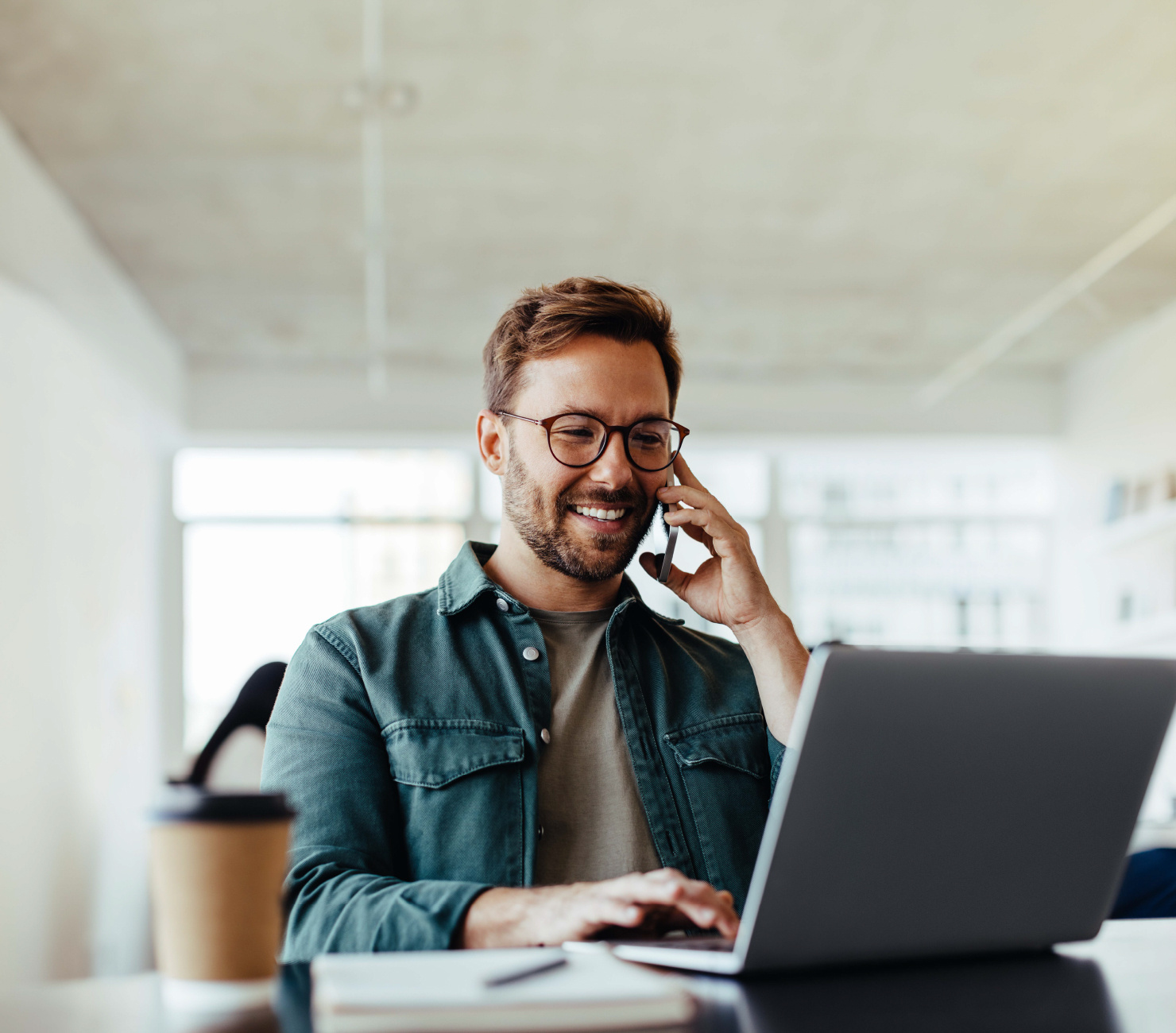 man sitting at a desk with his laptop open in front of him. He is on the phone and smiling