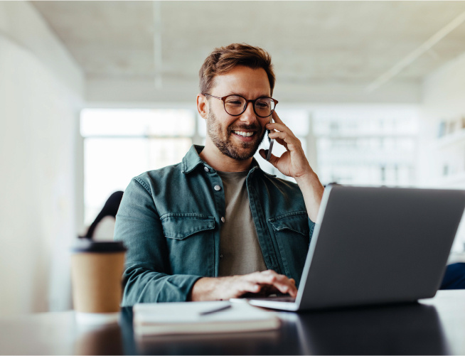 man sitting at a desk with his laptop open in front of him. He is on the phone and smiling. 