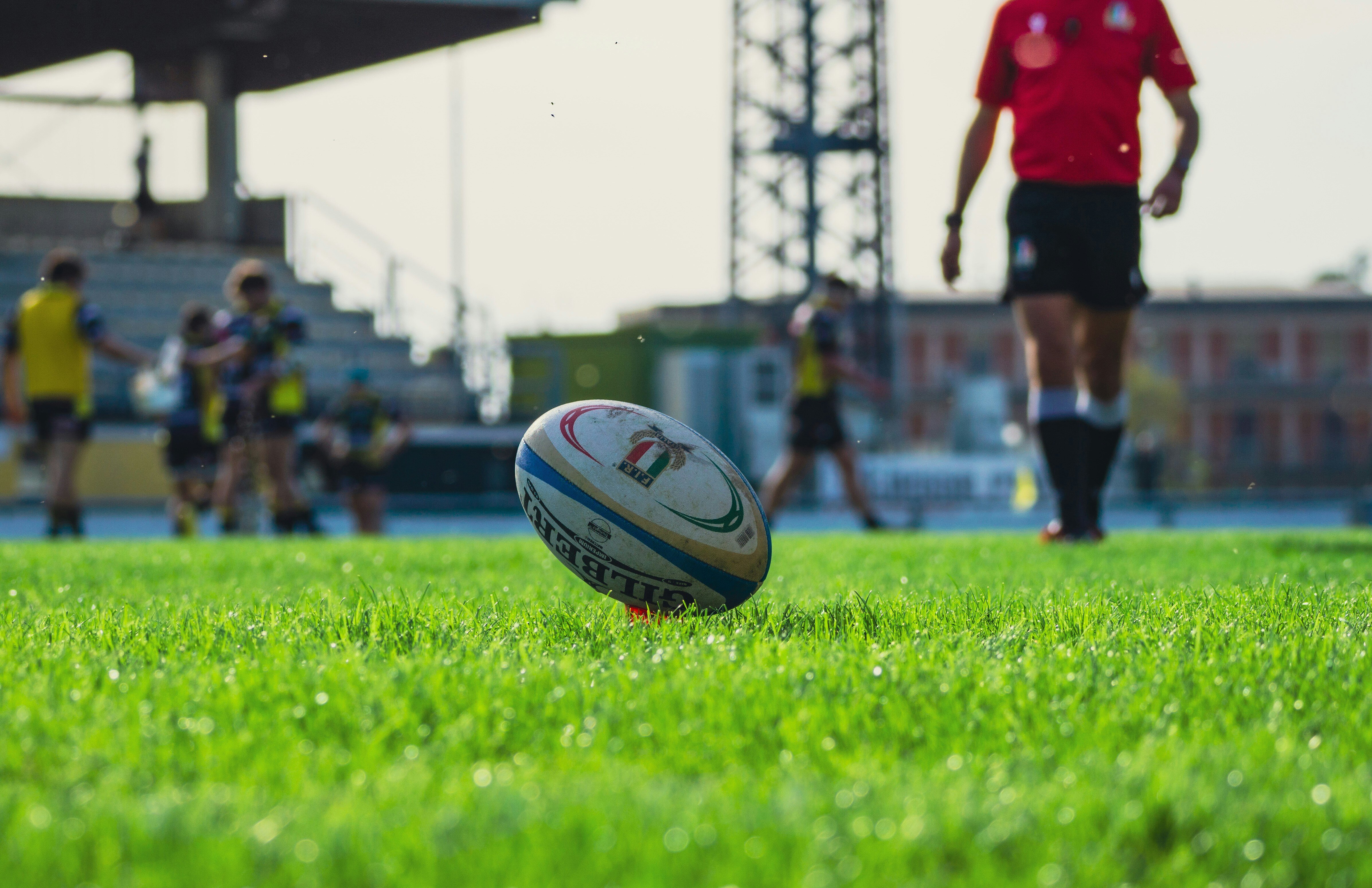 a man in a red rugby jersey walks towards a rugby ball on a green field